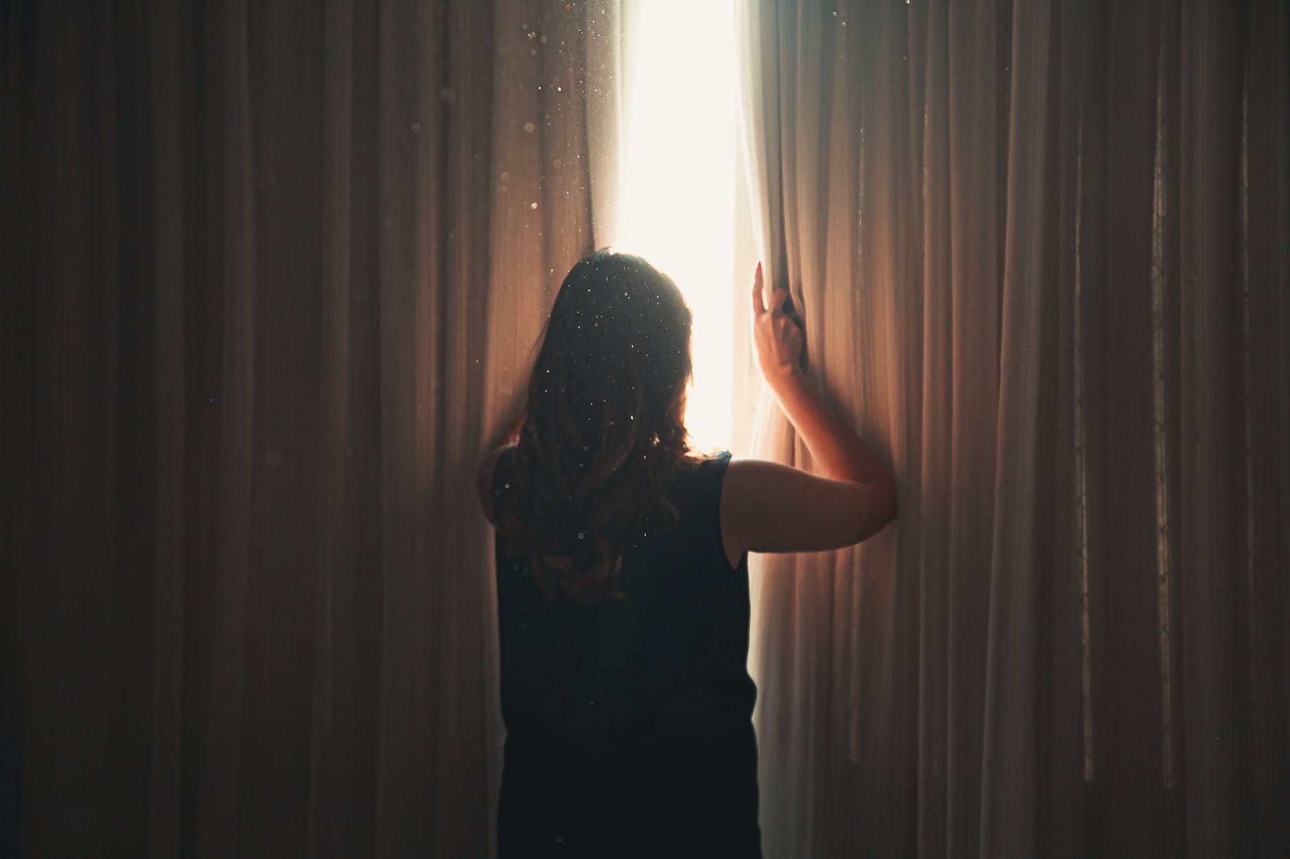 unrecognizable woman opening window curtain in shining daylight